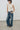 A model wearing a beige ruched sleeveless top and wide-leg blue denim jeans, standing against a plain light gray wall and concrete floor.