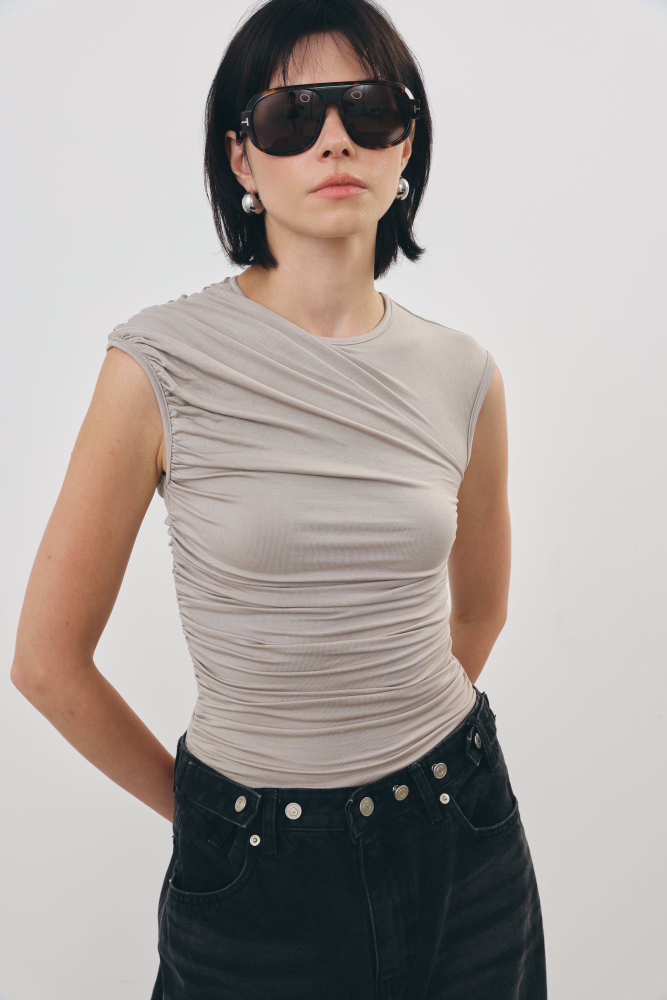 A woman wearing a sleeveless, ruched grey top and black jeans with silver button details, styled with dark sunglasses, against a plain light gray background.