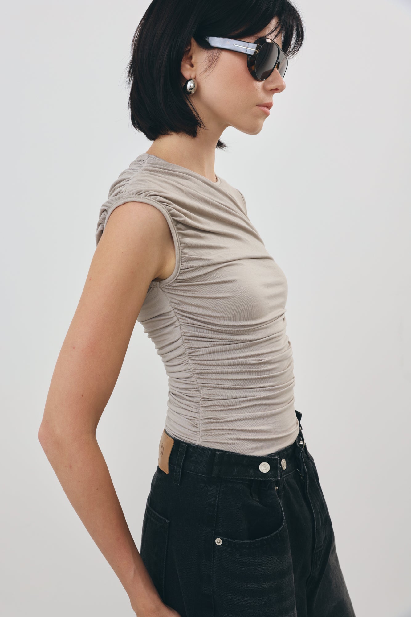 A woman in profile wearing a grey ruched sleeveless top and black jeans against a plain light gray studio background.