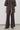 A model wearing a matching dark brown two-piece outfit consisting of a short-sleeve button-front top and wide-leg trousers with a cream drawstring, paired with dark loafers against a minimalist studio background.