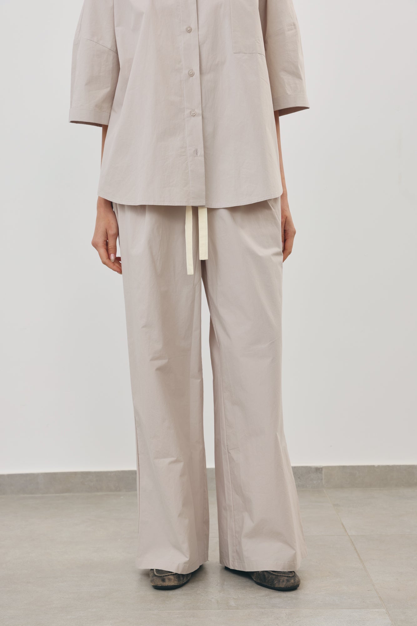 A person wearing a light beige matching two-piece outfit with a short-sleeve button-front top and wide-leg pants, standing against a plain light gray wall and floor.