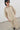 A model wearing an oversized beige short-sleeve button-up shirt and matching wide-leg trousers, styled with dark sunglasses and black shoes, standing in a minimalist room with a white wall and gray floor.