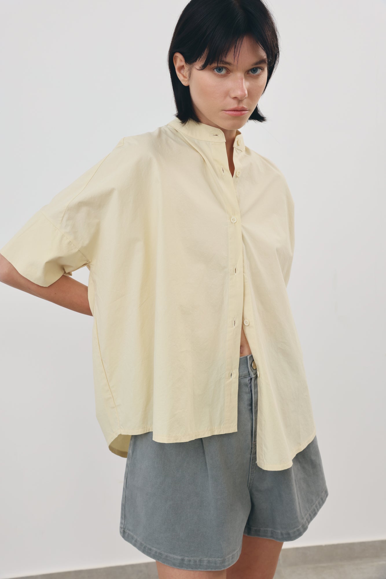 A model wearing an oversized, short-sleeved cream-colored button-up shirt and gray denim shorts, standing against a plain light gray wall.