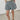 A person wearing high-waisted, wide-leg denim shorts in a faded light blue wash, paired with a cream shirt and dark loafers, standing against a plain light gray wall and floor.