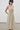 A woman modeling a saga midi dress with thin spaghetti straps, a smocked bodice, and a gathered skirt, set against a minimalist gray wall and tiled floor.