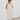 A woman with short black hair stands against a white wall wearing a cream-colored linen slip dress with lace trim on the straps and square neckline, paired with dark loafers.
