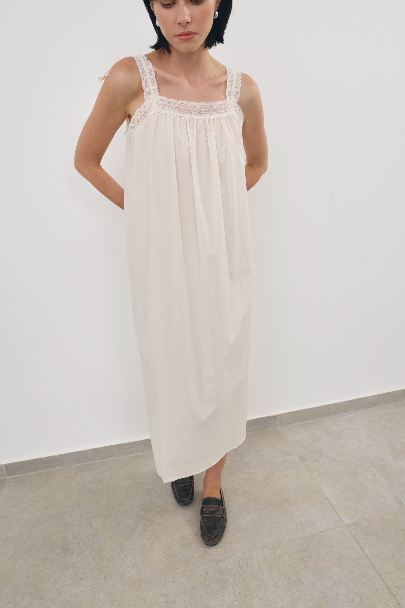 A woman wearing a cream-colored sleeveless maxi dress with lace trim on the straps, standing against a plain white wall.