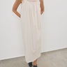 A woman wearing a cream-colored sleeveless maxi dress with lace trim on the straps, standing against a plain white wall.