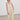 A model wearing a cream-colored viscosa midi dress with a smocked bodice and thin lace shoulder straps, paired with khaki flat shoes, standing in a minimalist studio setting.