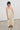 A model wearing a cream-colored viscosa midi dress with a smocked bodice and thin lace shoulder straps, paired with khaki flat shoes, standing in a minimalist studio setting.