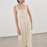 A model wearing a cream-colored viscosa midi dress with a smocked bodice and thin lace shoulder straps, paired with khaki flat shoes, standing in a minimalist studio setting.