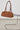 A brown suede duffle bag with dual top handles, hanging from a clear acrylic chair with a purple cushion on a metal cantilever chair in a minimalist room.