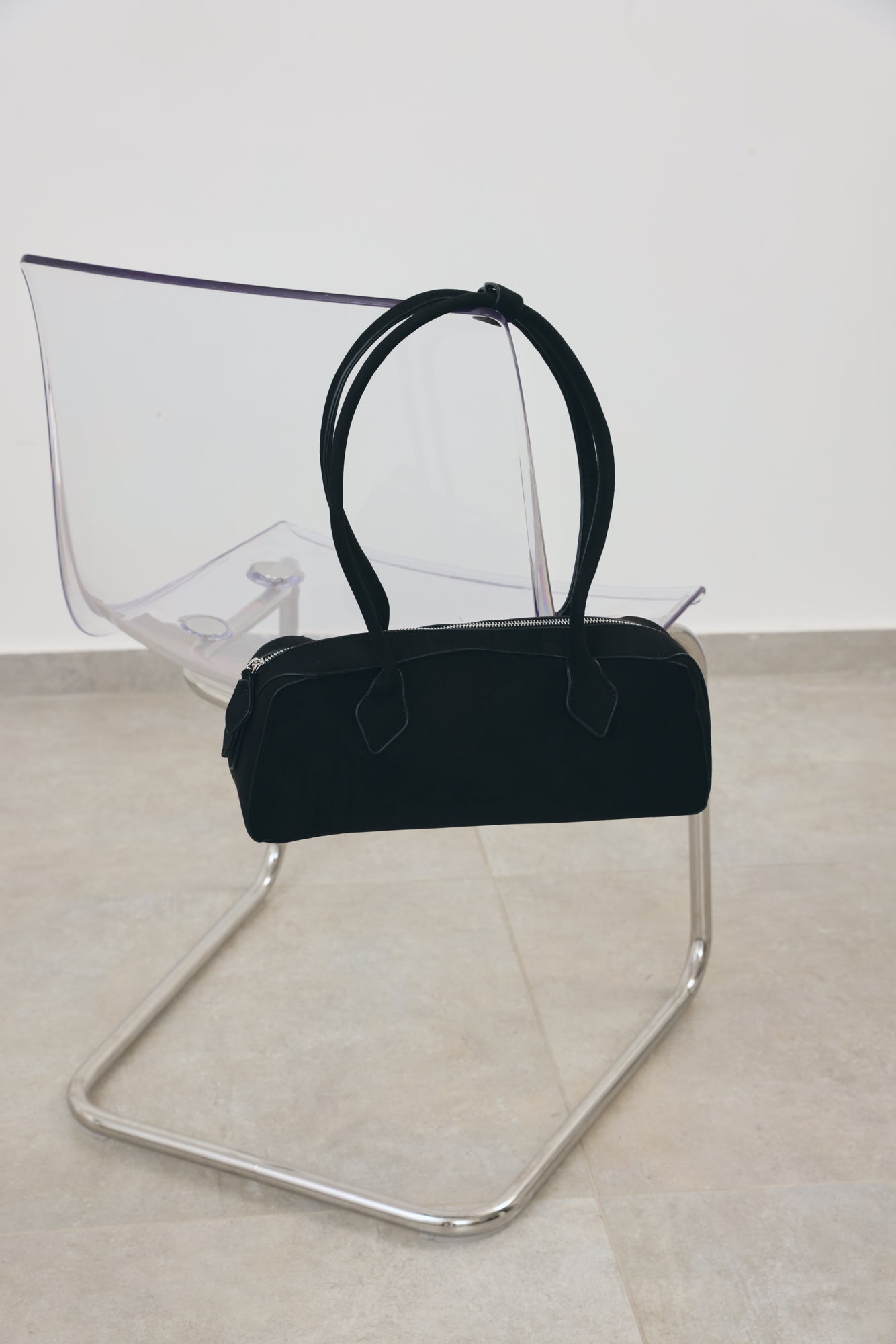 A black fabric handbag with double top handles, displayed on a clear acrylic and chrome chair against a minimalist off-white wall and light beige floor.
