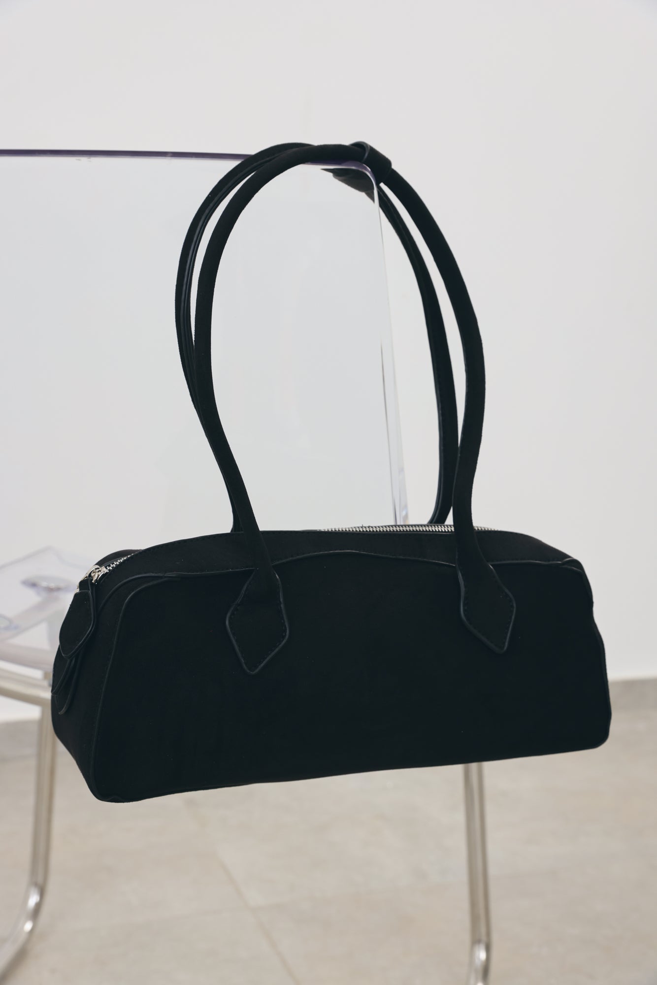 A black fabric duffel-style handbag with two long shoulder straps, displayed hanging from a clear stand in a minimalist indoor setting.